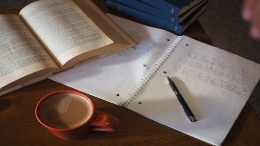 A photo of a tabletop with a stack of books, open notebook with handwritten notes and a pen, and a cup of coffee. 