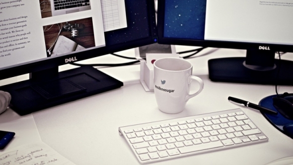 A photograph of a desk with two monitors, a keyboard, a coffee mug, and papers.