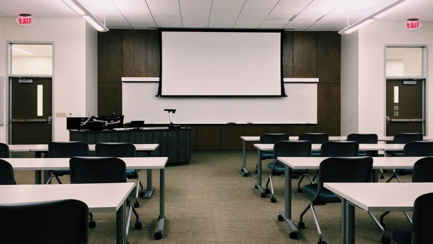 A photograph of an empty classroom with a projector at the front of the classroom and rows of two-seated desks. 