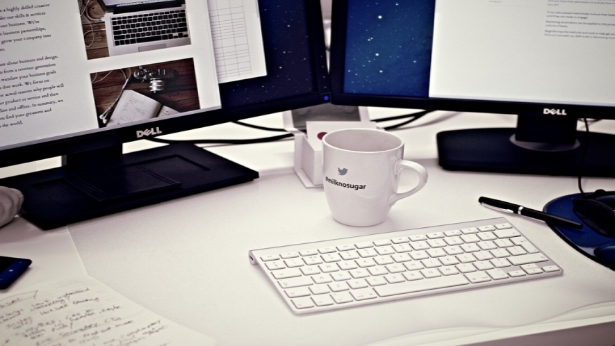 A photograph of a desk with two monitors, a keyboard, a coffee mug, and papers.