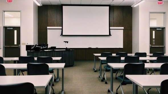 A photograph of an empty classroom with a projector at the front of the classroom and rows of two-seated desks. 